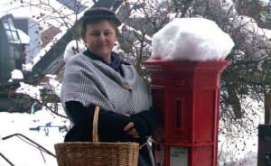 A woman dressed in period victorian clothing, holding a picnic basket, standing beside a red postbox which is covered in snow